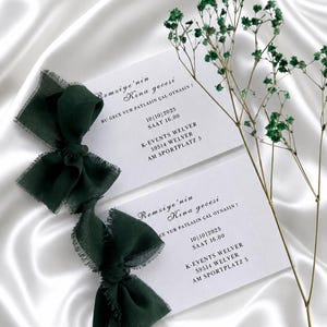 May include: Two white invitation cards with dark green text and a dark green ribbon bow. The cards have event details, including a date and time. Green sprigs are placed near the cards on a white satin surface.
