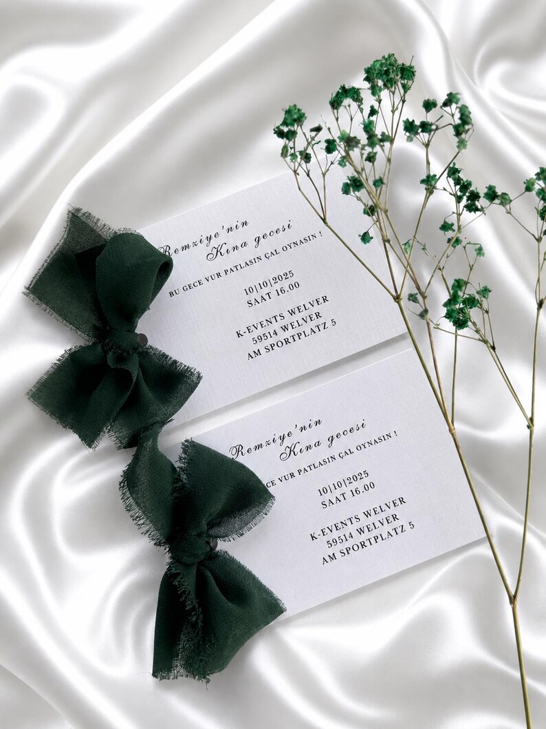 May include: Two white invitation cards with dark green text and a dark green ribbon bow. The cards have event details, including a date and time. Green sprigs are placed near the cards on a white satin surface.