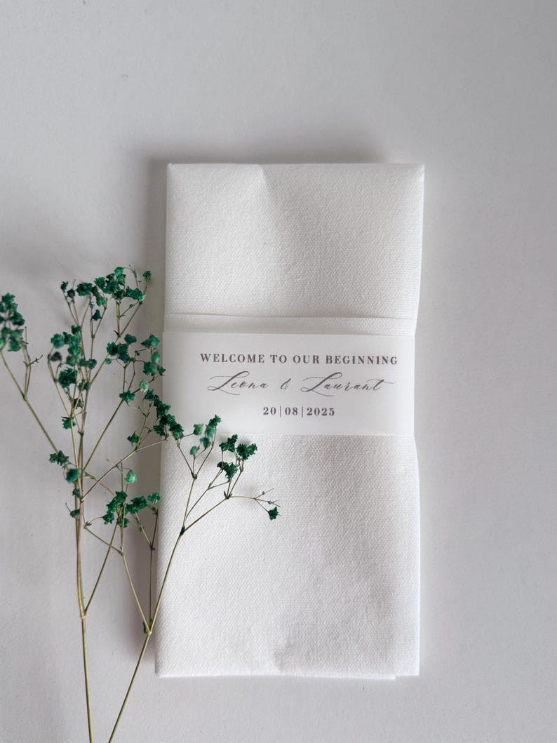 May include: A folded white napkin with a band that reads "WELCOME TO OUR BEGINNING" with the names Leena & Laurent and the date 20|08|2025. Green sprigs of dried flowers are on the left side of the napkin.