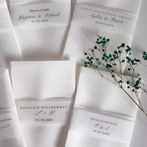 May include: White folded napkins with translucent bands bearing personalized text and dates. The napkins are arranged with a sprig of green foliage. The text includes names and dates, suggesting a wedding or special event.