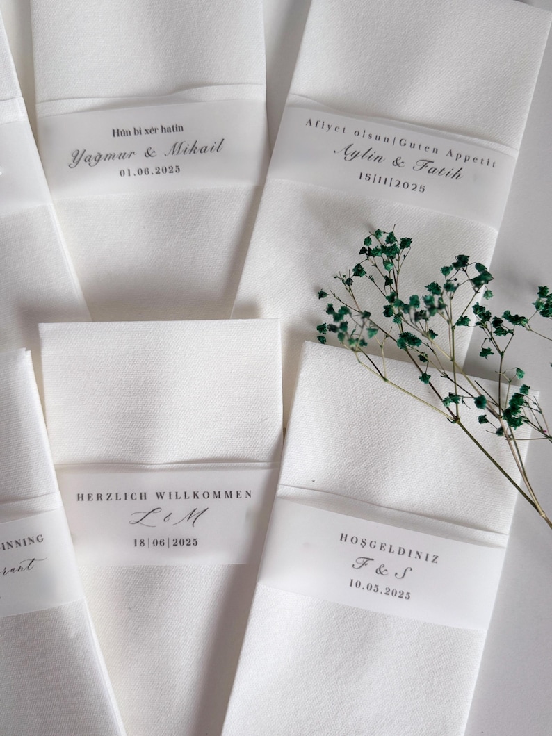 May include: White folded napkins with translucent bands bearing personalized text and dates. The napkins are arranged with a sprig of green foliage. The text includes names and dates, suggesting a wedding or special event.
