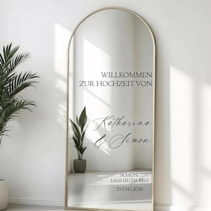 Könnte beinhalten: Ein gewölbter Standspiegel mit einem dünnen goldenen Rahmen und schwarzer Schrift: WILLKOMMEN ZUR HOCHZEIT VON Katharina & Simon SCHON, DASS DU DA BIST. 19|04|2026. Der Spiegel reflektiert eine Topfpflanze und einen hellen, neutralen Innenraum.