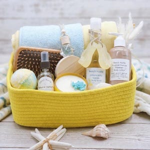 May include: A yellow woven basket filled with spa products. Includes blue and yellow towels, a hairbrush, bath bombs, body wash, body lotion, a candle, and a natural sunspray. The products have the text "Beach Hustle" on them.