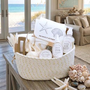 May include: A white rope basket filled with beach-themed spa products. The basket contains a variety of items, including bottles, soaps, and a card with a starfish and seashell design. The text on the card reads "the beach is my happy."