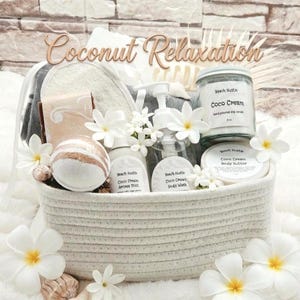 May include: A white woven basket containing a collection of bath products including a bath bomb, Coco Cream body butter, Coco Cream body wash, and Coco Cream aroma mist. The set features white plumeria flowers, seashells, and a gray towel.