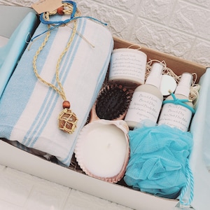 May include: A white gift box filled with a variety of spa and bath products, including a blue and white striped towel, a seashell candle, a wooden hair brush, a blue bath pouf, a bottle of sea salt bath mist, a bottle of natural body wash, and a brown stone necklace with a wooden bead.