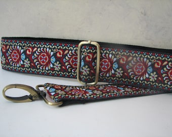 May include: Black bag strap with a repeating floral pattern in red, blue, and gold. The strap has gold-coloured metal hardware, including a buckle and a swivel clasp. The strap is designed to be adjustable and attachable to a bag.