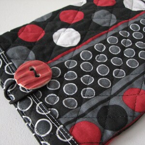 May include: A quilted fabric item featuring a black base with a pattern of red, white, and grey polka dots and circles. A red button with two holes fastens the item. Likely a case or cover.