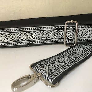 May include: Black guitar strap with a silver floral design. The strap has a silver buckle and a silver hook.