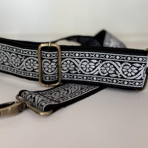 May include: Black and silver patterned bag strap with gold-colored metal hardware. The strap features a repeating floral design in silver against a black background. Includes a rectangular buckle and a swivel clasp.