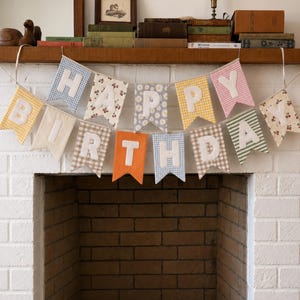 Peut inclure: Une banderole en tissu épelant HAPPY BIRTHDAY avec des fanions individuels aux motifs et couleurs variés, incluant du vichy, des fleurs et de l'orange uni. Chaque fanion présente une lettre en tissu blanc cousue au centre, suspendue par une fine corde.