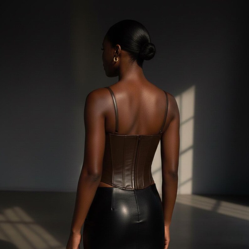 May include: A brown faux leather corset top with thin straps and a zipper closure. The top is paired with a black leather skirt. The model is facing away from the camera, with their hair pulled back.