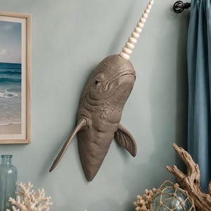 May include: A wall-mounted narwhal sculpture featuring a textured gray body with visible skin folds and a long, spiraled white tusk extending upward from the head. The marine mammal figure includes two side fins and a detailed eye.