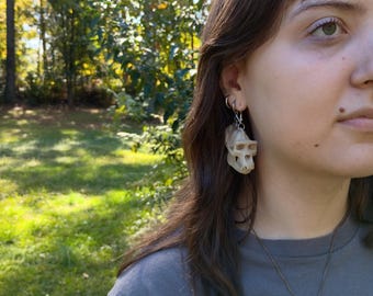 May include: A dangling earring featuring a small, off-white, realistic primate skull replica. The skull is suspended from a silver-toned metal hook and jump ring, showcasing detailed bone structure, including eye sockets, a nasal cavity, and teeth.