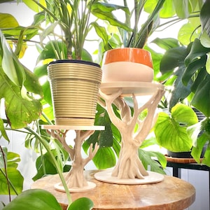 以下が含まれることがあります： 幹のデザインが施された2つのプラントスタンドが、鉢植えの植物を支えています。ポットは金色とオレンジと白。スタンドは丸い木製のテーブルの上にあり、緑豊かな葉に囲まれています。