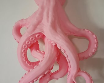 May include: A wall-mounted octopus sculpture in a solid light pink color. The creature features a rounded head with two prominent, almond-shaped yellow-green eyes and eight textured tentacles that curve and extend outward from the central body.