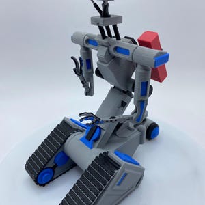 Puede incluir: Un juguete robot de color gris y azul impreso en 3D con un ladrillo rojo en la espalda. El robot tiene una cabeza grande con dos ojos azules y un cuerpo gris con detalles azules. El robot está sentado sobre una superficie blanca.