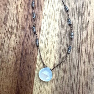 Könnte beinhalten: Eine zarte Halskette mit einem hellblauen, facettierten Tropfenanhänger. Die braune Kordel ist mit kleinen, grauen Perlen verziert. Der Anhänger ist der Mittelpunkt und fängt das Licht durch seine vielen Facetten ein.