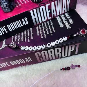 Puede incluir: Una pila de libros con los títulos "Hideaway" y "Corrupt" está coronada por figuritas de calaveras y un marcapáginas de cuentas. El marcapáginas deletrea "Devils Night" con cuentas de letras blancas y presenta un dije de cruz.