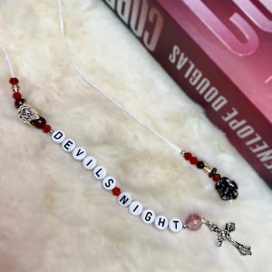Puede incluir: Un marcapáginas de cuerda blanca con cuentas rojas y transparentes, un dije plateado y cuentas de letras blancas que deletrean "DEVILS NIGHT".