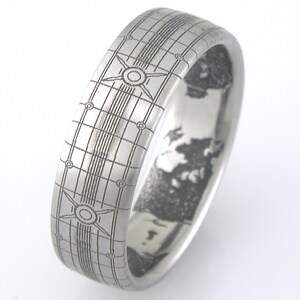 May include: A silver ring with a grid pattern and a circular design on the top. The inside of the ring has a detailed engraved design.