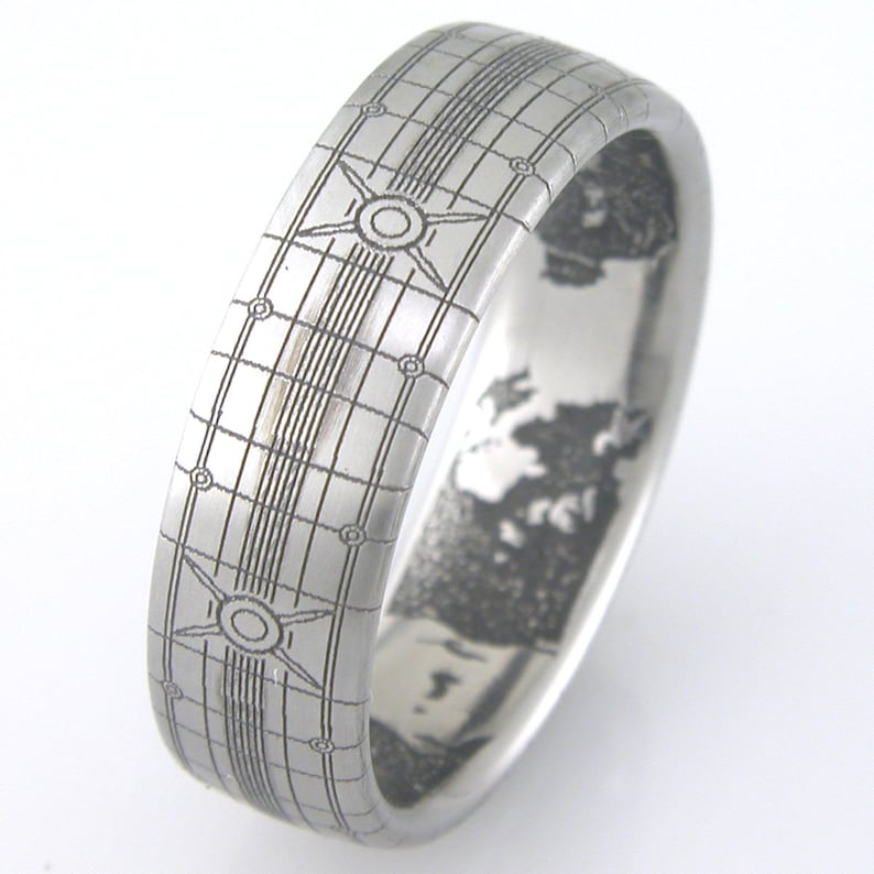 May include: A silver ring with a grid pattern and a circular design on the top. The inside of the ring has a detailed engraved design.