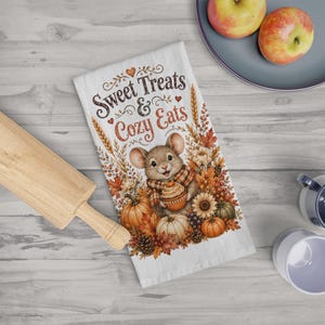 Puede incluir: Un paño de cocina blanco con una ilustración central de un ratón con bufanda de cuadros que sostiene un cupcake. El texto dice Sweet Treats & Cozy Eats, rodeado de motivos otoñales que incluyen calabazas, girasoles y espigas de trigo.