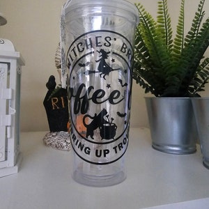 Peut inclure: Un gobelet en plastique transparent avec un motif noir et blanc qui dit "Witches Brew Coffee Stirring Up Trouble". Le motif représente une sorcière sur un balai, un chat noir et un chaudron.
