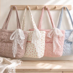 May include: Four quilted tote bags hanging on a wooden rack. The bags feature gingham patterns in brown, pink, and blue, or a white fabric with small red hearts. Each bag is personalised with embroidered names and adorned with a white lace bow.