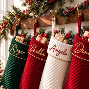 Puede incluir: Cuatro calcetines navideños de terciopelo acanalado en verde oscuro, rojo, blanco y rojo. Cada calcetín tiene nombres bordados en oro o rojo: Roger, Bella, Angela y Dina. Están rellenos con pequeñas cajas de regalo, ramas de pino y bayas.