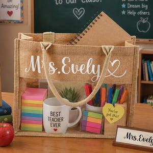 Może przedstawiać: Jutowa torba z napisem "Mrs. Evely" w białym skrypcie, wypełniona przyborami szkolnymi. Zawiera kubek z napisem "Best Teacher Ever", karteczki samoprzylepne, długopisy i notatnik. Widoczny jest r&oacute;wnież drewniany szyld z napisem "Mrs. Evely".