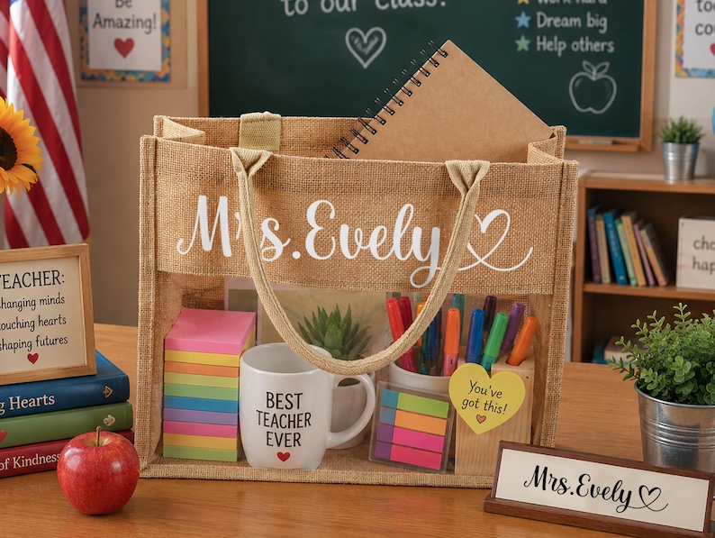 Może przedstawiać: Jutowa torba z napisem "Mrs. Evely" w białym skrypcie, wypełniona przyborami szkolnymi. Zawiera kubek z napisem "Best Teacher Ever", karteczki samoprzylepne, długopisy i notatnik. Widoczny jest r&oacute;wnież drewniany szyld z napisem "Mrs. Evely".