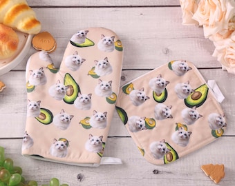 May include: A light pink oven mitt and pot holder set. The set features a repeating pattern of white cats and avocado slices. The oven mitt is a standard size, and the pot holder is square.