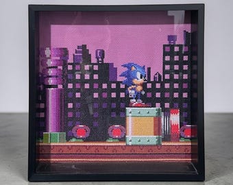 May include: Cross-stitch artwork of Sonic the Hedgehog in a framed shadow box. The scene depicts a pixelated cityscape with a purple sky and green and brown foreground. The artwork is in a black frame.