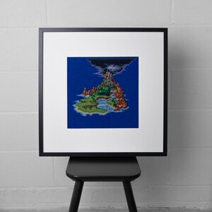 May include: A square framed cross-stitch embroidery featuring a pixel art landscape of a tropical island with a central mountain, a dark storm cloud at the peak, green vegetation, and blue water, all set against a solid deep blue background.