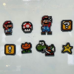 May include: A selection of pixel art designs featuring characters and items from the Super Mario video game. The set includes Mario, a mushroom, a question mark block, a flower, Yoshi, and a star, in various colours.