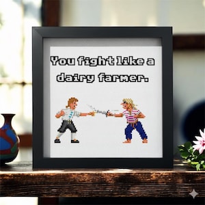 May include: Black framed cross-stitch art with the text "You fight like a dairy farmer." Two pixelated figures are depicted fencing. The artwork is displayed on a wooden surface with decorative items.