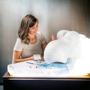 May include: A person is painting a white plaster cast of a pregnant belly. The cast is sitting on a table with a light underneath it. There are paintbrushes and tubes of paint on the table.