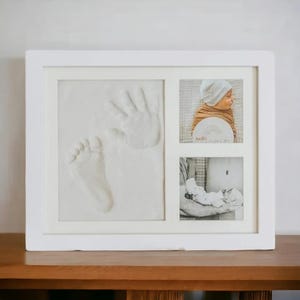 May include: White picture frame with three openings. The top opening shows a baby's handprint and footprint in white plaster. The middle opening shows a baby in a white hat and swaddled in a blanket. The bottom opening shows a baby being held by a person. The text "hello world" is visible in the middle opening.