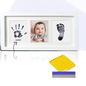 May include: White wooden frame with three openings for photos. The top opening has a black handprint with the name "HARPER" underneath. The middle opening has a color photo of a baby. The bottom opening has a black footprint. The frame comes with a yellow ink pad and a blue scraper.