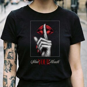 Könnte beinhalten: Schwarzes T-Shirt mit einer Grafik einer roten Lippe und einem Finger vor dem Mund, mit dem Text "Shut Your Mouth". Das Design ist in einem schwarzen Rechteck gerahmt. Das Model steht auf einer Stadtstraße.
