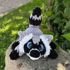 Op de afbeelding: Een gehaakte wasbeerknuffel met een grijs en wit lichaam en zwarte accenten rond de ogen, oren en pootjes. Het knuffeldier heeft grote, ronde, felblauwe plastic ogen en een opvallende, gestreepte staart die omhoog staat.