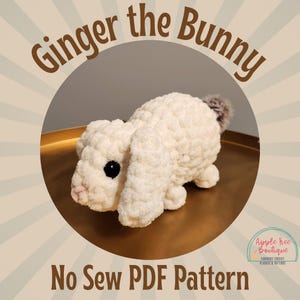 Op de afbeelding: Een wit gehaakt konijn genaamd Ginger, met zwarte ogen en een roze neus. Het konijn zit op een gouden oppervlak. De tekst "Ginger the Bunny No Sew PDF Pattern" staat onderaan de afbeelding. De tekst "Apple Tree Boutique Handmade Crochet Plushies & Patterns" staat rechtsonder.