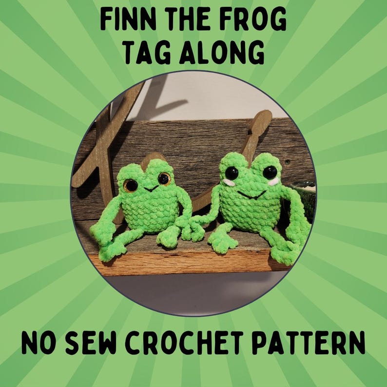 May include: Two green crocheted frog plushies with black eyes and smiling mouths. The frogs are sitting on a wooden surface. The text "Finn the Frog Tag Along No Sew Crochet Pattern" is displayed above the frogs.