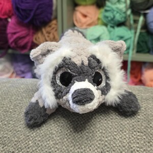 Op de afbeelding: Een gehaakte wasbeer knuffel gemaakt van zacht grijs, wit en donker antraciet garen. Het knuffeldier heeft grote, ronde, zwart glinsterende ogen, een witte snuit, een donkere neus en pluizige witte vachtaccenten rond het gezicht en de oren.