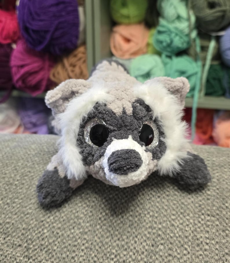 Op de afbeelding: Een gehaakte wasbeer knuffel gemaakt van zacht grijs, wit en donker antraciet garen. Het knuffeldier heeft grote, ronde, zwart glinsterende ogen, een witte snuit, een donkere neus en pluizige witte vachtaccenten rond het gezicht en de oren.