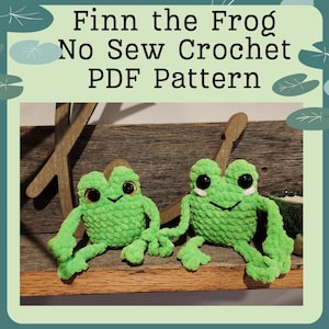 May include: Two green crocheted frog plushies with black eyes and white smiles sitting on a wooden surface. The text "Finn the Frog No Sew Crochet PDF Pattern" is displayed above the frogs.