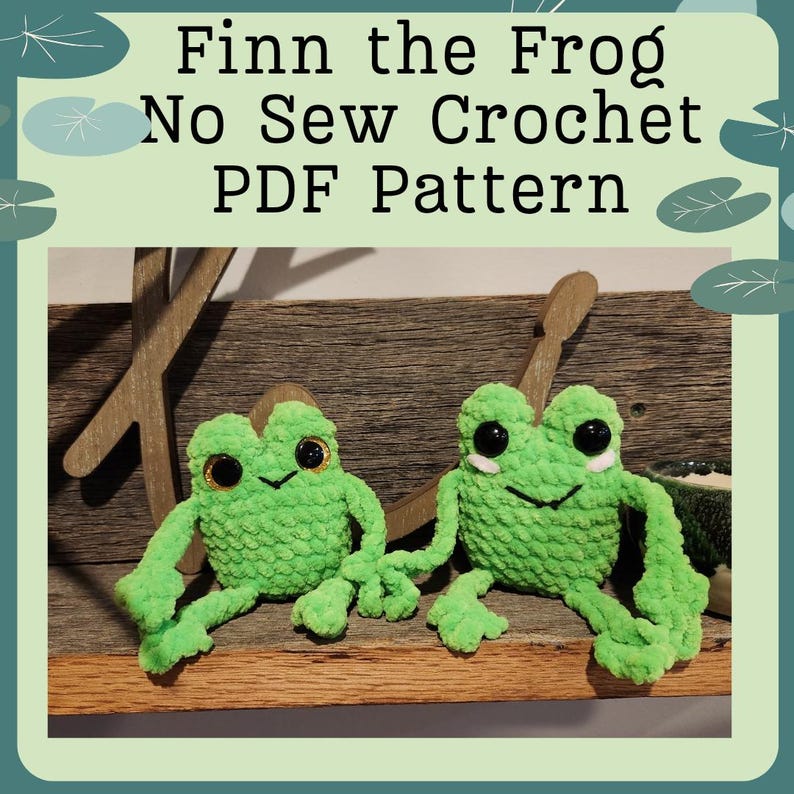 May include: Two green crocheted frog plushies with black eyes and white smiles sitting on a wooden surface. The text "Finn the Frog No Sew Crochet PDF Pattern" is displayed above the frogs.