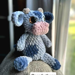 May include: A blue and gray crocheted cow plush toy with pink nose and sparkly brown eyes. The toy is sitting on a gray surface. @comfortloops
