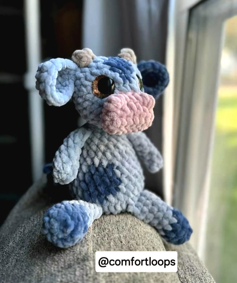 May include: A blue and gray crocheted cow plush toy with pink nose and sparkly brown eyes. The toy is sitting on a gray surface. @comfortloops